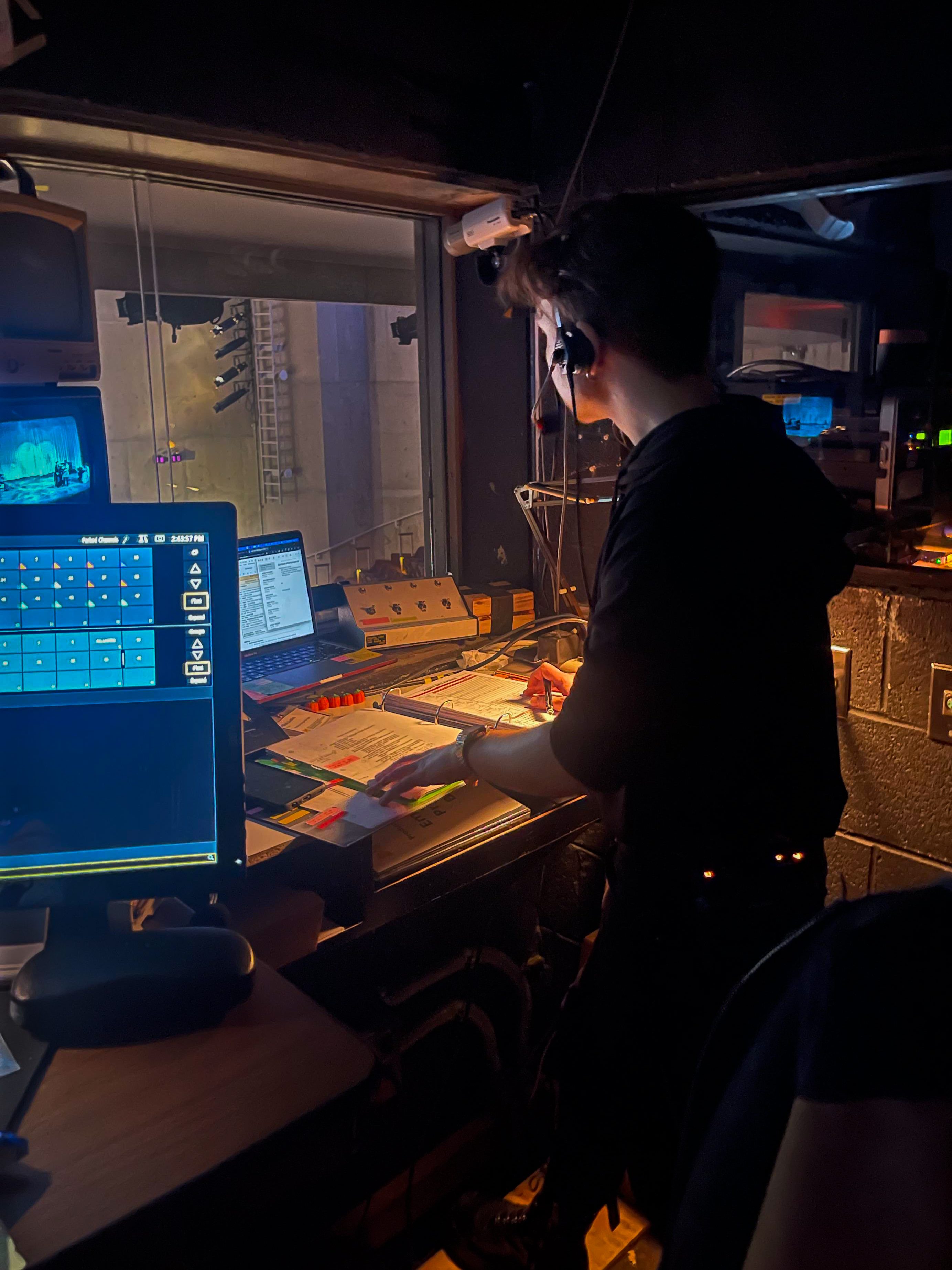 A photo of Miles Zoellick in a booth, calling a show. He is wearing a headset and has his hands on his call script, a laptop in front of him, a cue light switchboard to his right, and two monitors to his left.