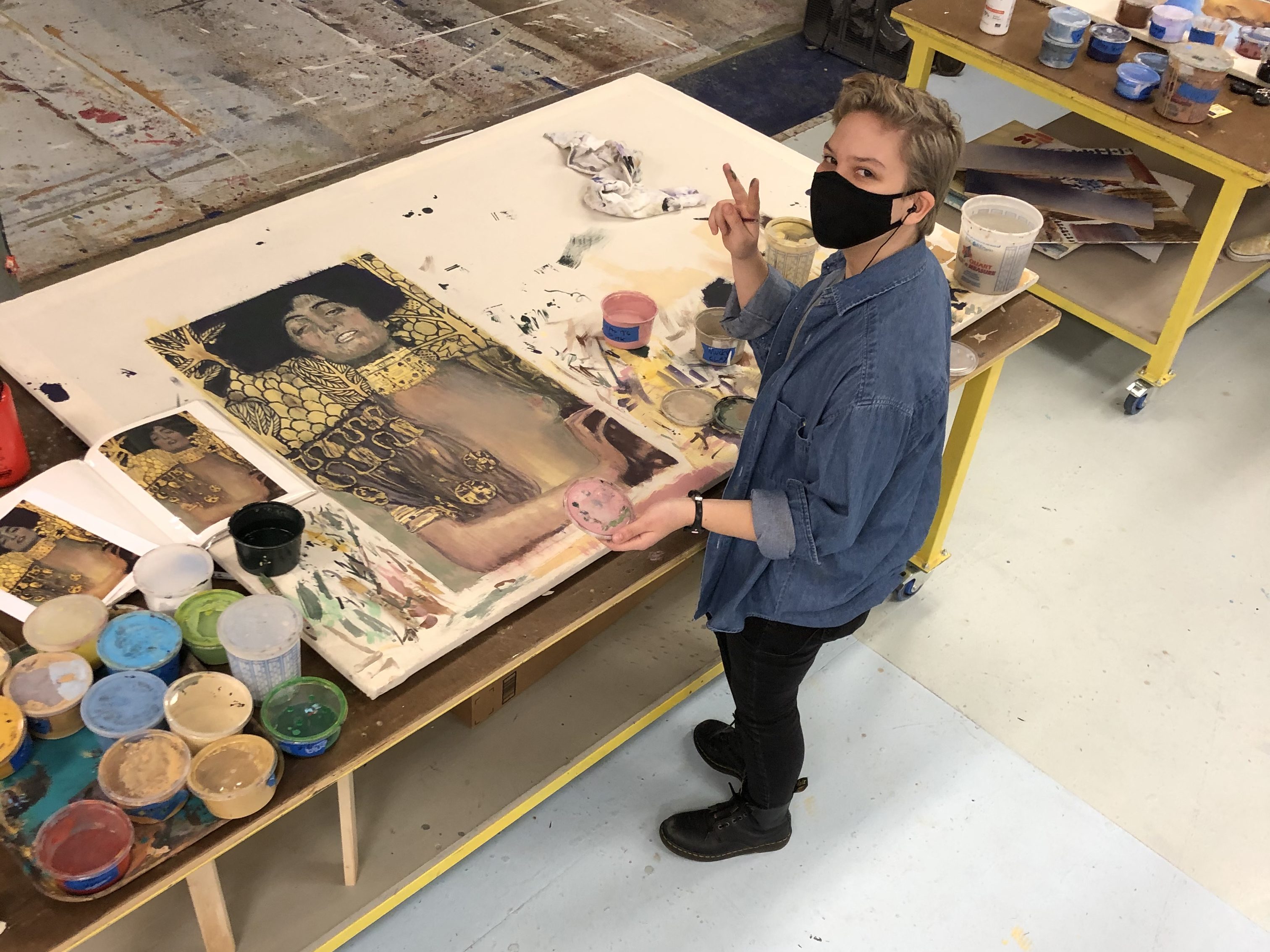 A photo of Miles in the process of painting a study of Gustav Klimt's Judith I. He is wearing a black face mask and holding up a peace sign.