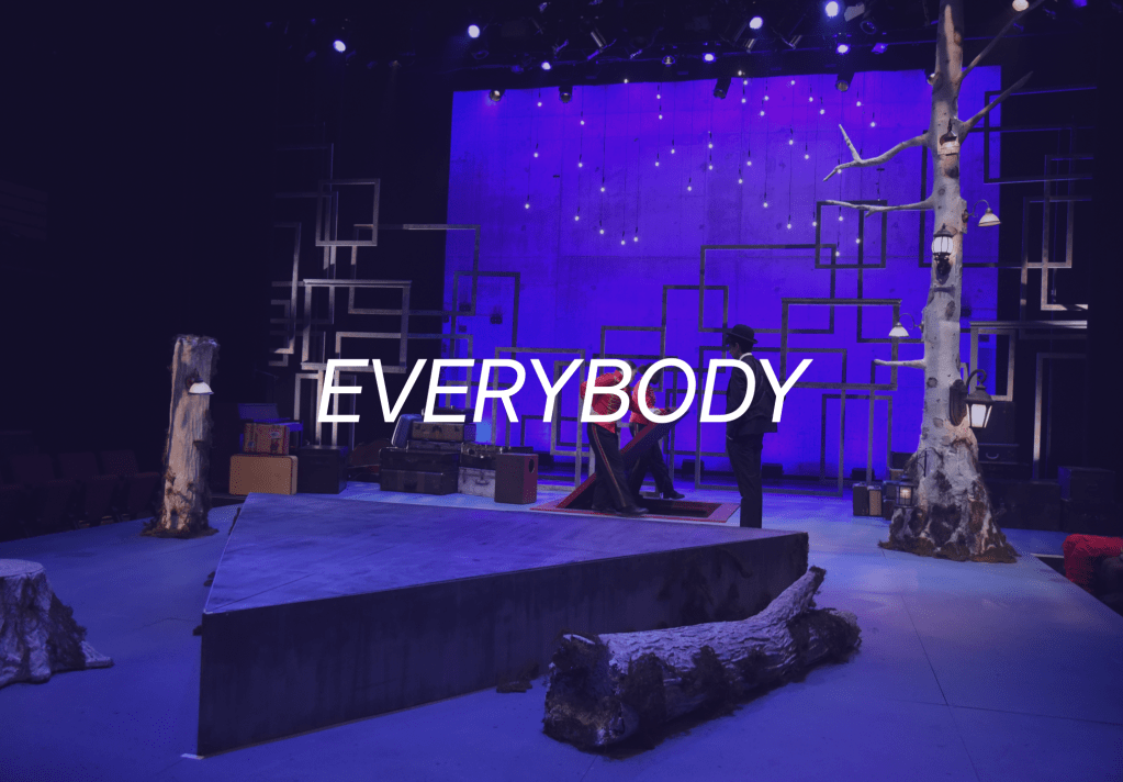 Everybody (Cover Image: An eclectic set featuring a diamond platform, felled trees, floating frames, and hanging bulbs under purple lighting. Two performers upstage open a red trap door.)